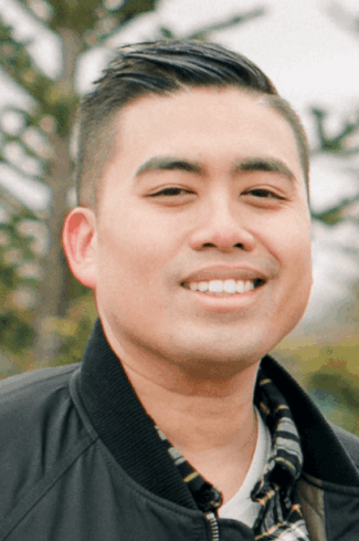 Portrait of a smiling man with short hair, wearing a black jacket and plaid shirt, outdoors with soft-focus trees in the background.
