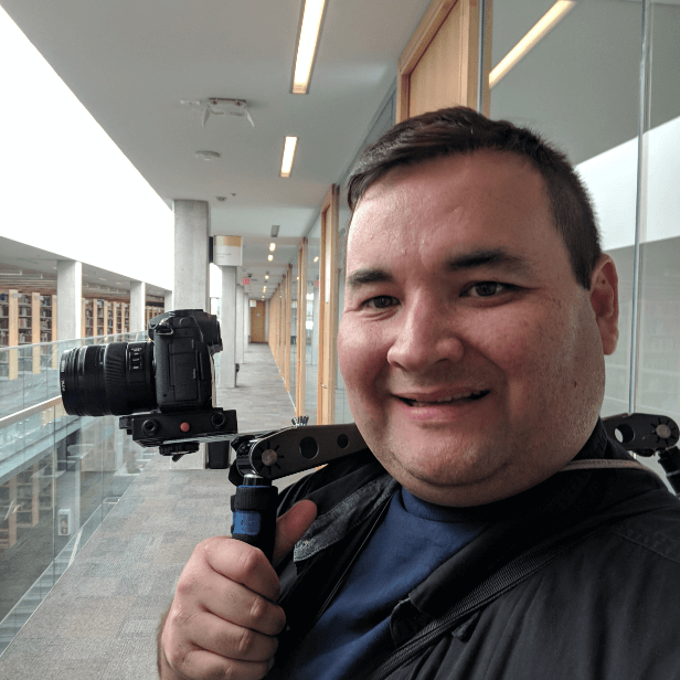 Man smiling while holding a camera on a gimbal in a brightly lit hallway with large windows.