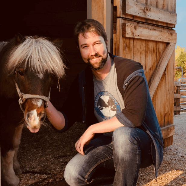 Man in a denim jacket kneeling next to a pony outside a wooden stable, smiling at the camera during sunset.
